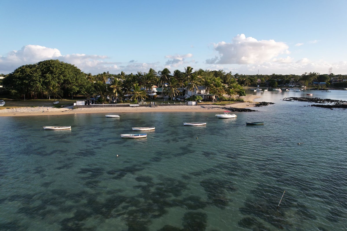 A serene view of the beach is presented, showcasing several small boats anchored in calm waters. Lush palm trees frame the shoreline, while the soft sand leads to a residential area in the background, providing a relaxing coastal atmosphere.