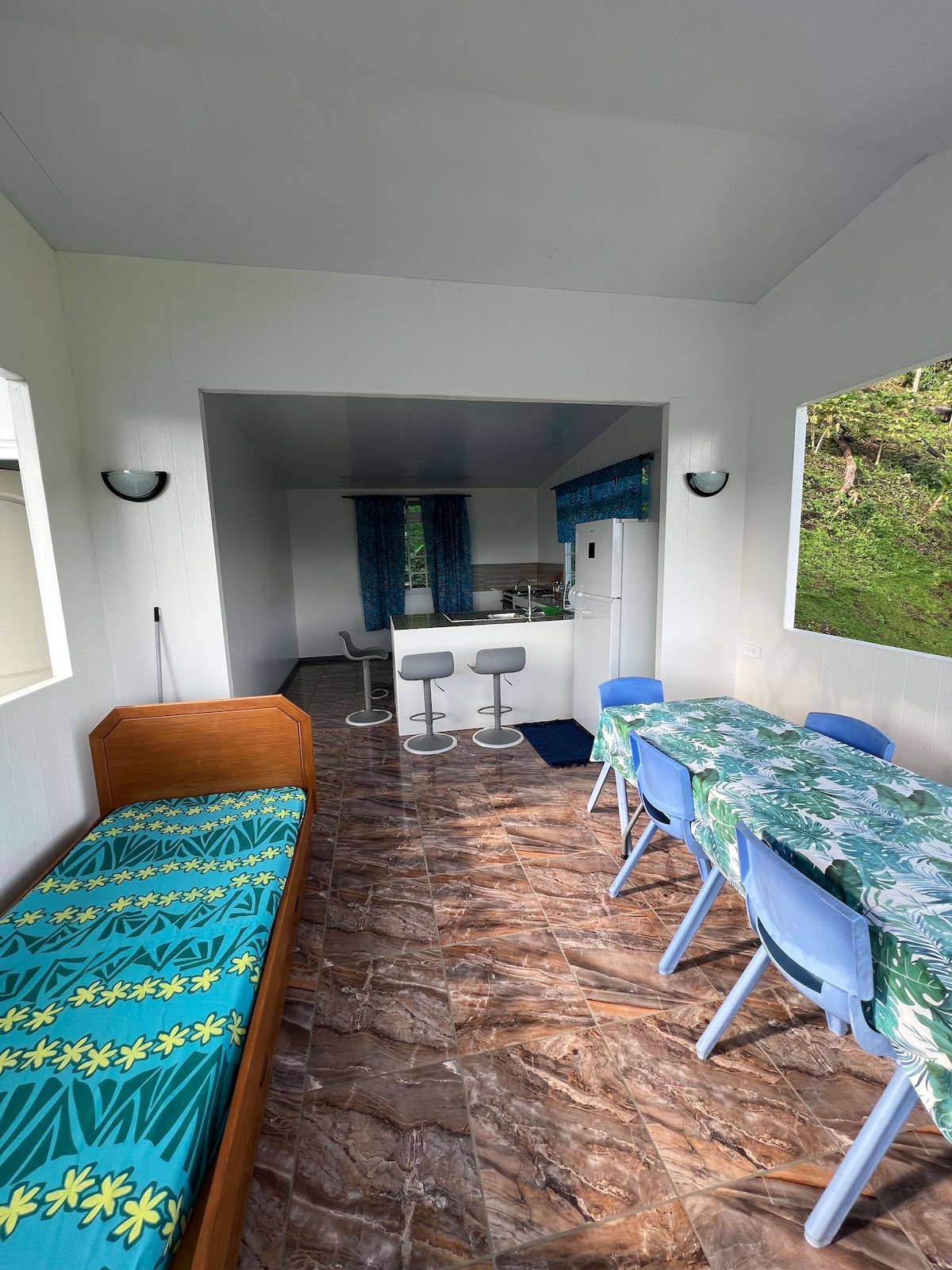 An open living space features a light-colored tiled floor and large windows allowing natural light. A dining table set with blue chairs is situated beside a small sofa, while two bar stools are positioned at a kitchen counter in the background.