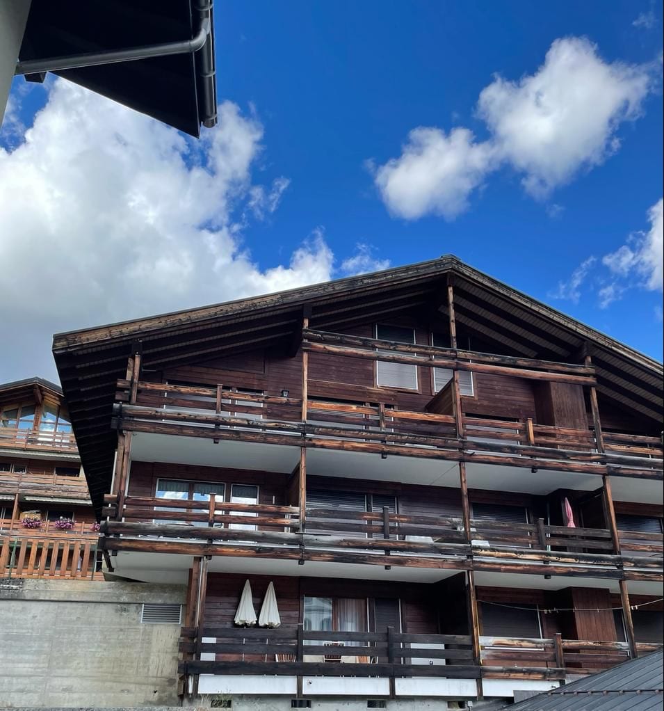 A wooden chalet-style building is displayed against a bright blue sky with scattered clouds. Multiple balconies with wooden railings extend from the facade, providing outdoor space. The architecture features a sloped roof and a combination of large windows and rustic elements.