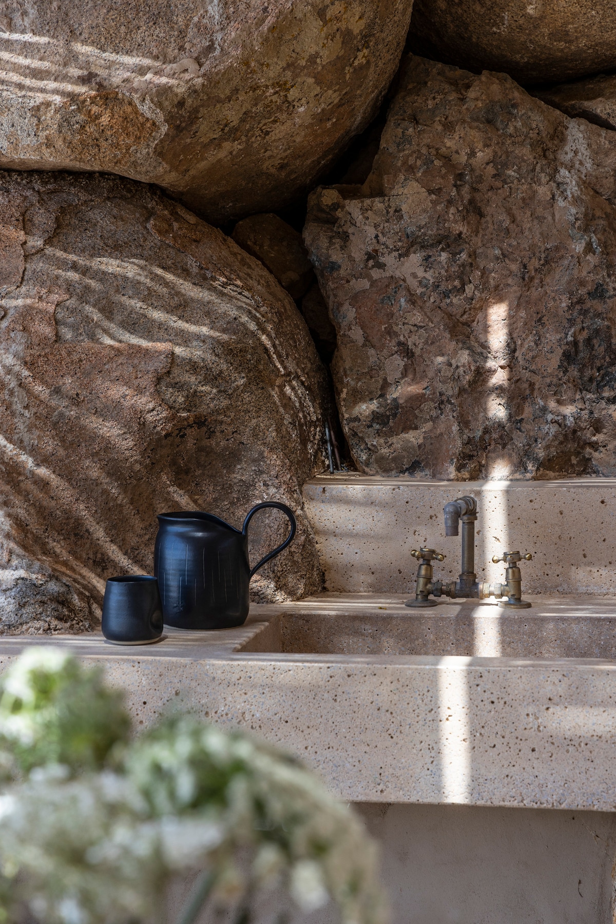 A minimalist stone sink is set against a backdrop of large, rugged boulders. A sleek black pitcher and a cup are positioned on the countertop, with light filtering through nearby elements, creating a gentle play of shadows.