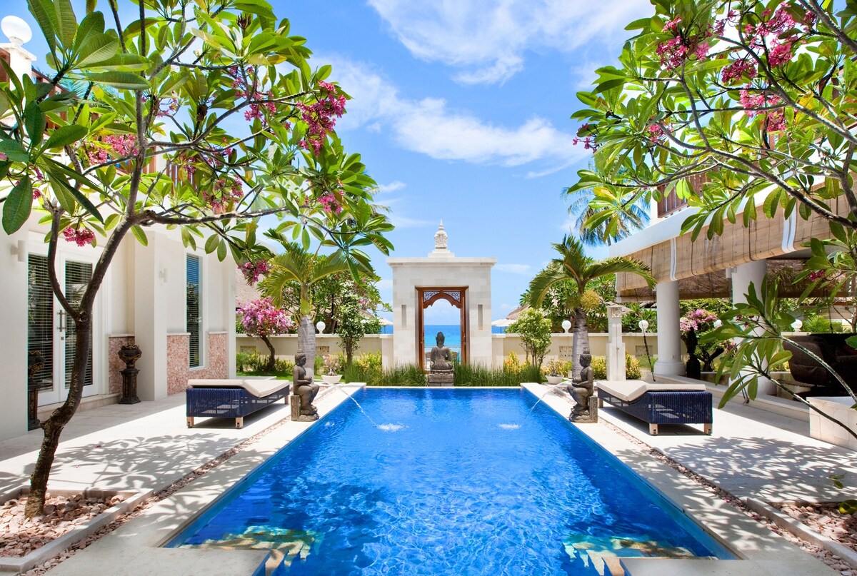 A serene outdoor area features a vibrant blue pool surrounded by lush tropical plants and blooming flowers. Relaxing lounge chairs are positioned at the poolside. An elegant structure with a decorative entrance can be seen in the background, framed by greenery leading to an expansive view of the sky.
