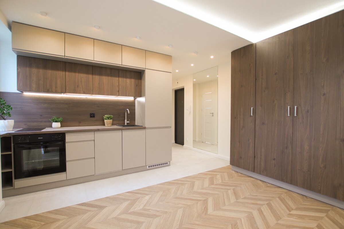 A modern kitchen space features sleek cabinetry in neutral tones, complemented by warm wood accents. A built-in oven and stylish countertop are visible, along with a plant adding a touch of greenery. The floor displays a herringbone pattern, enhancing the room's contemporary feel.