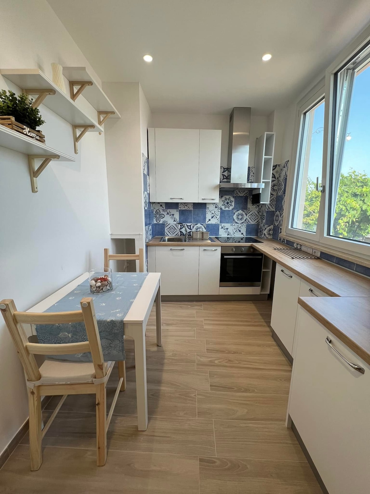 A modern kitchen with a clean design is showcased. It features white cabinetry and blue tiled backsplash, complemented by natural light from large windows. A small table with two chairs is positioned near the window, providing a cozy dining area.