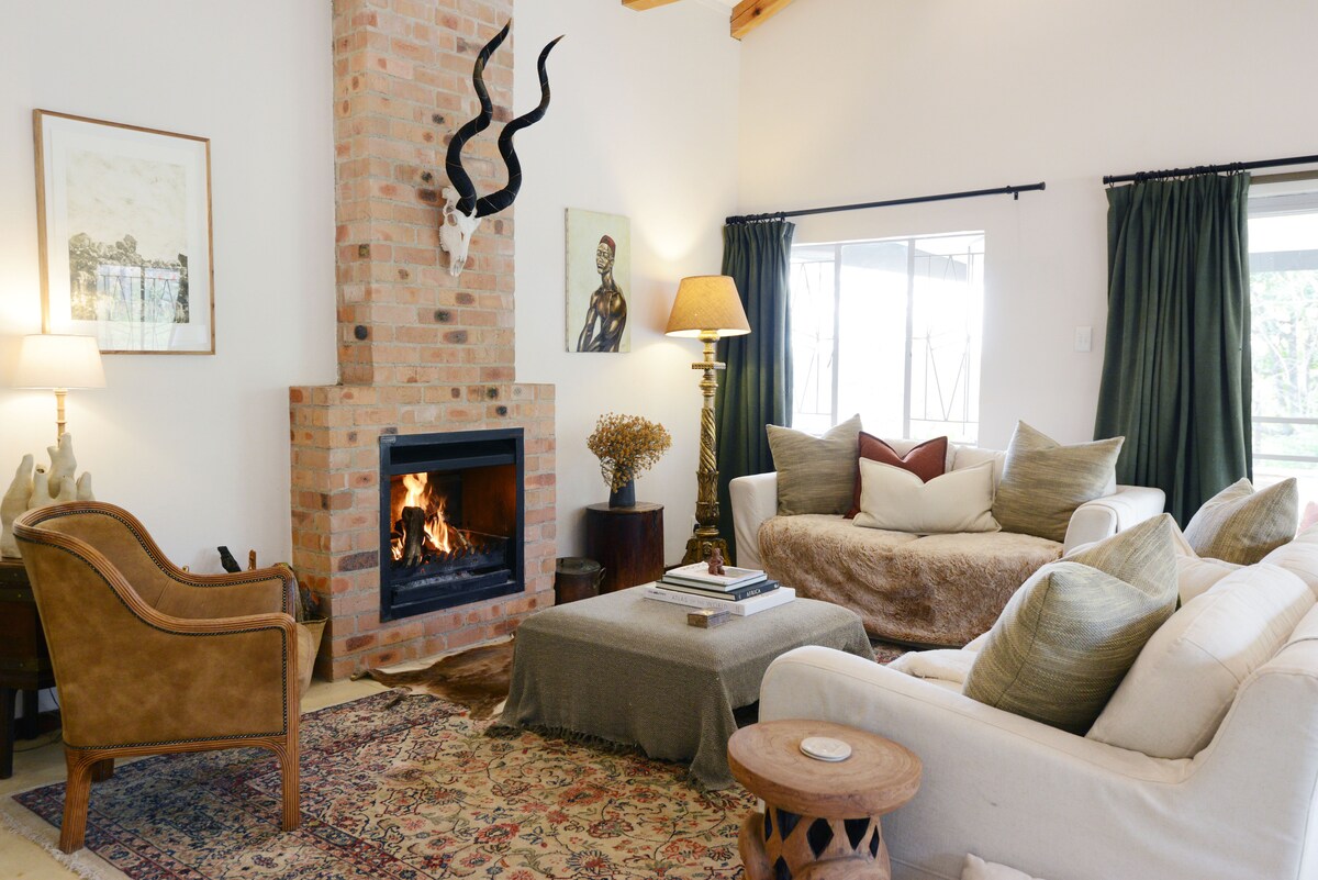 A comfortable living area features a brick fireplace with a mounted animal trophy above it. Plush seating options include a large sofa adorned with various cushions, complemented by a cozy throw. A textured area rug adds warmth, while natural light enters through the nearby windows.