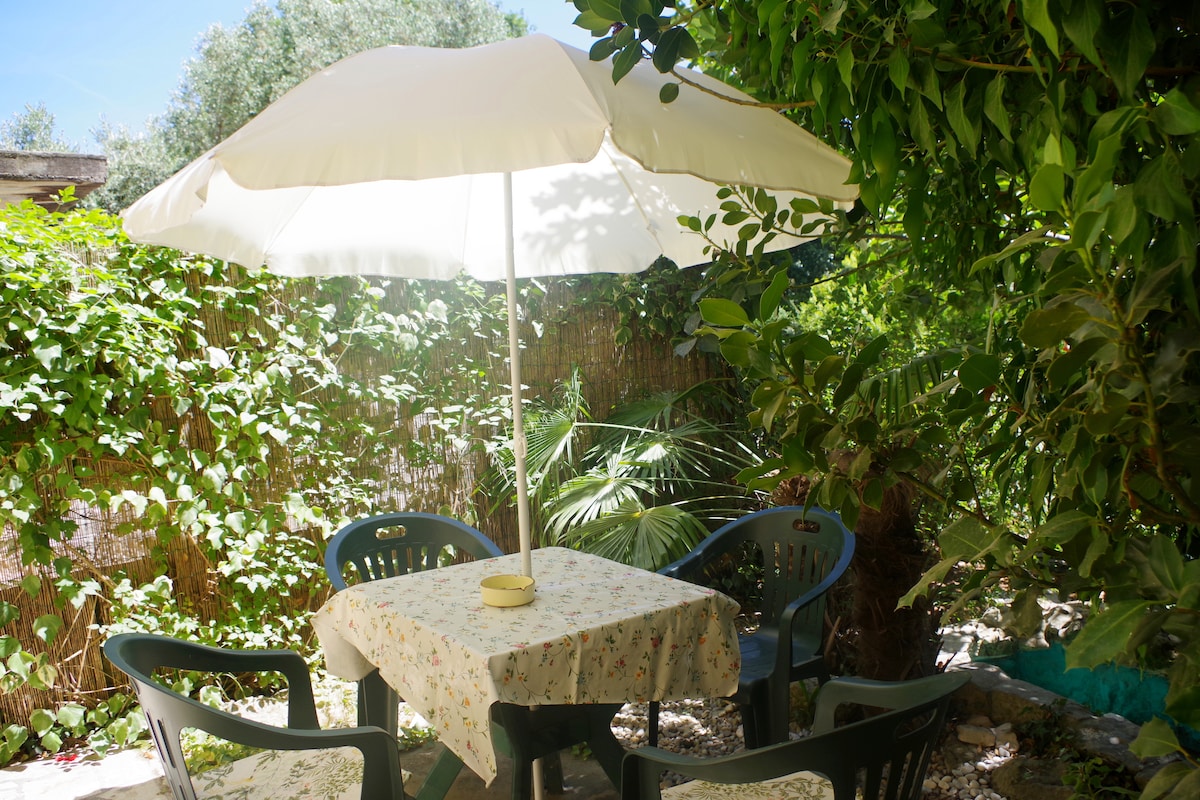 A shaded outdoor terrace is surrounded by lush greenery, featuring a table draped with a floral tablecloth and four black plastic chairs. A large umbrella provides protection from sunlight, offering a comfortable space for relaxation or dining.