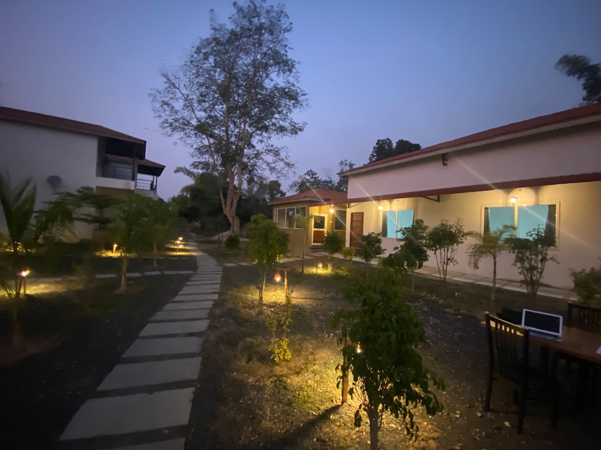 A tranquil pathway lined with soft lighting leads through the landscaped garden of the resort. Two buildings are visible, each with illuminated windows. Surrounding plants and trees provide a natural setting, enhancing the serene atmosphere as dusk settles.