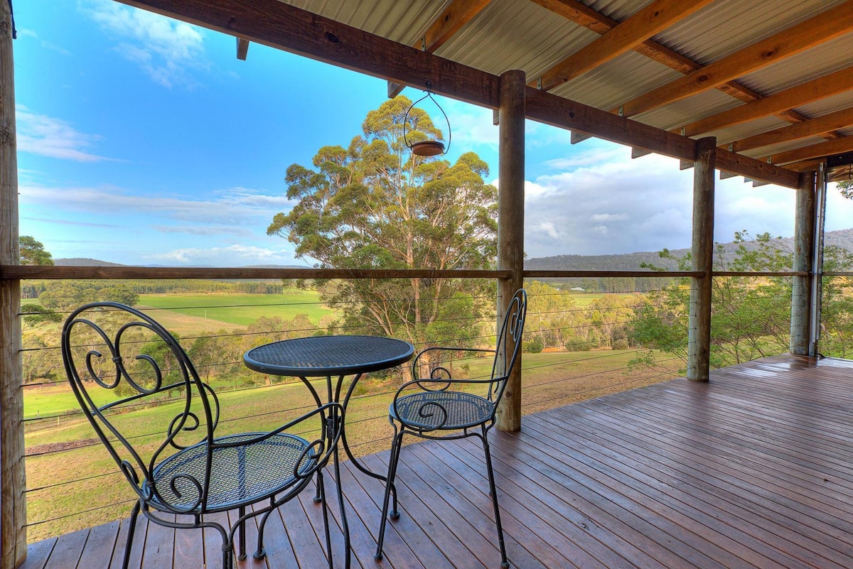 A covered balcony is presented with a small circular table and two wrought iron chairs. Natural wood beams and a polished wooden floor frame the outdoor space, which overlooks lush farmland and distant trees, creating a peaceful connection to the surrounding environment.