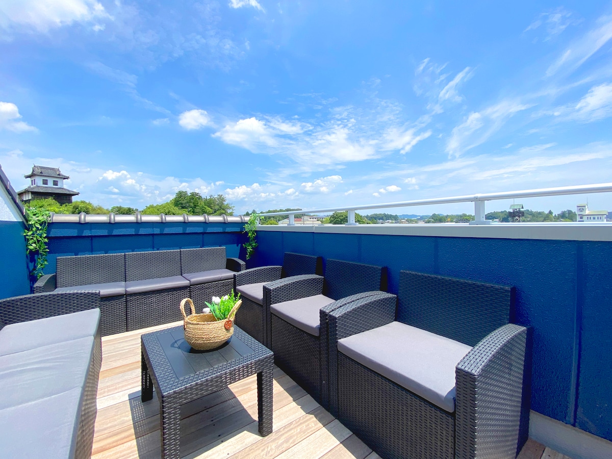 An outdoor terrace features comfortable seating arranged around a central table, providing a space for relaxation. The area is surrounded by a low blue wall, offering views of the sky and nearby landscape. Greenery adds a touch of nature to the setting.
