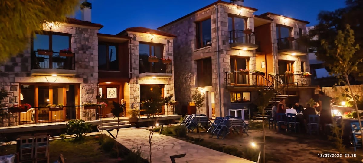 Two stone buildings are illuminated at night, showcasing spacious balconies with plants. A well-lit pathway leads to the entrance, surrounded by vibrant greenery and seating areas. The inviting atmosphere is enhanced by warm lighting, highlighting the rustic charm of the architecture.