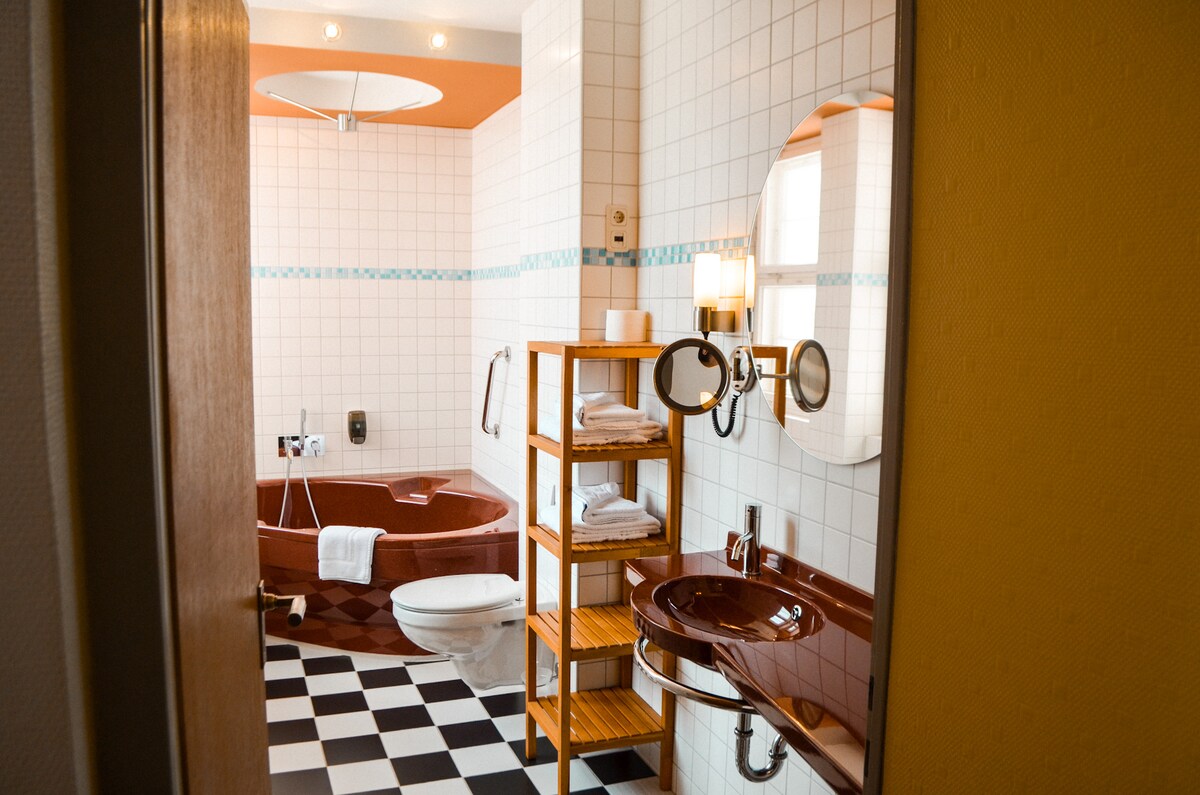 A brightly coloured bathroom features a round tub and a modern toilet. A wooden shelving unit holds neatly arranged towels. A circular mirror above a stylish sink complements the checkered floor, while natural light is provided by an overhead fixture.
