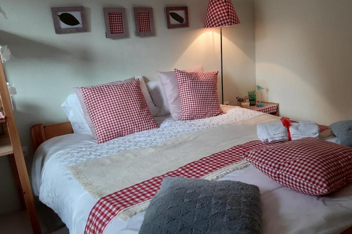 A cozy sleeping area features a spacious bed dressed in white linens with a patterned red-and-white checkered blanket. Two decorative pillows complement the bedding, while a bedside lamp casts a soft glow. A small side table holds various essential items and personal touches.