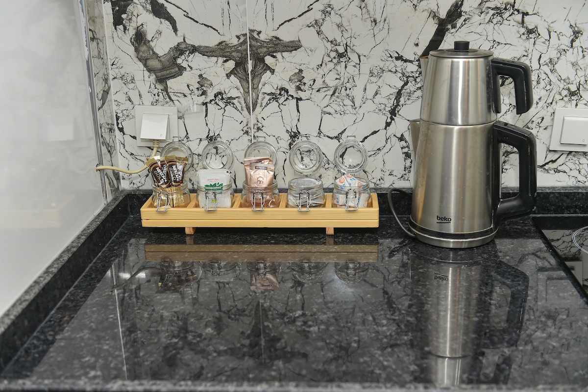 A sleek kitchen countertop is presented with a polished black surface. A row of decorative glass cups rests on a wooden tray, with a silver kettle positioned nearby. The backdrop features a striking marble wall design that adds depth to the space.