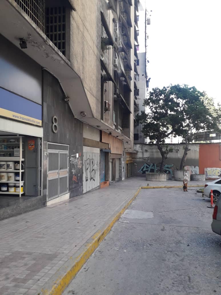 A city street scene is depicted, featuring a multi-story building with a smooth facade. Graffiti adorns the walls, and a narrow pathway leads towards the entrance. Sparse vegetation is visible beside the street, with parked vehicles in view.