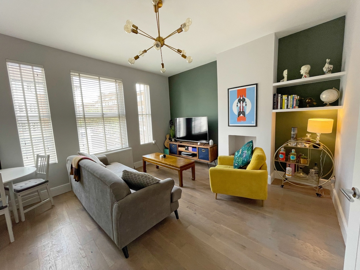 A well-lit living room features large windows with white blinds, allowing natural light to fill the space. A comfortable gray sofa and a bright yellow accent chair are positioned around a wooden coffee table. A television is mounted on the wall, with shelves displaying decorative items.