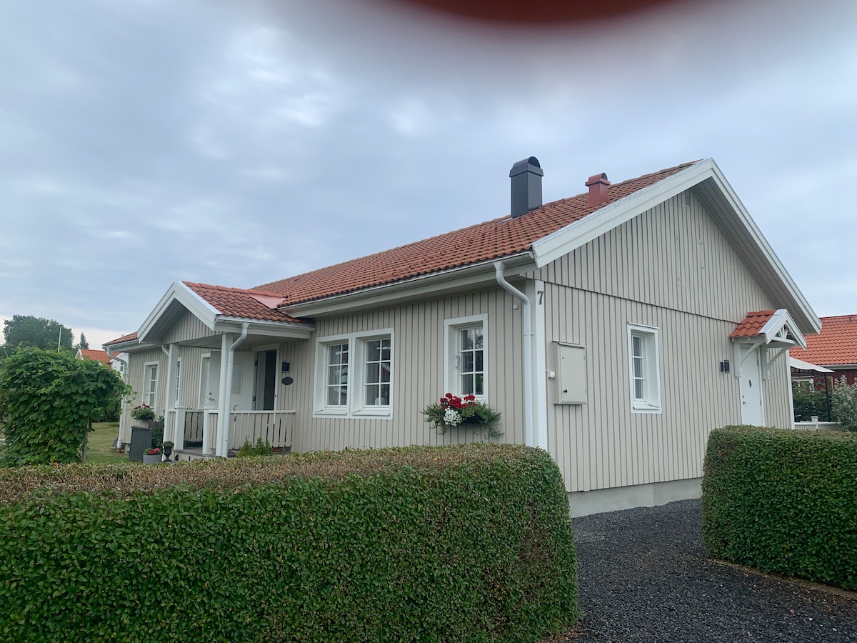 A single-story house is framed by neatly trimmed hedges and a gravel pathway. The light-colored wooden exterior features a red-tiled roof, and large windows allow ample natural light. Planters with flowers are positioned by the entrance, enhancing the home's inviting presence.