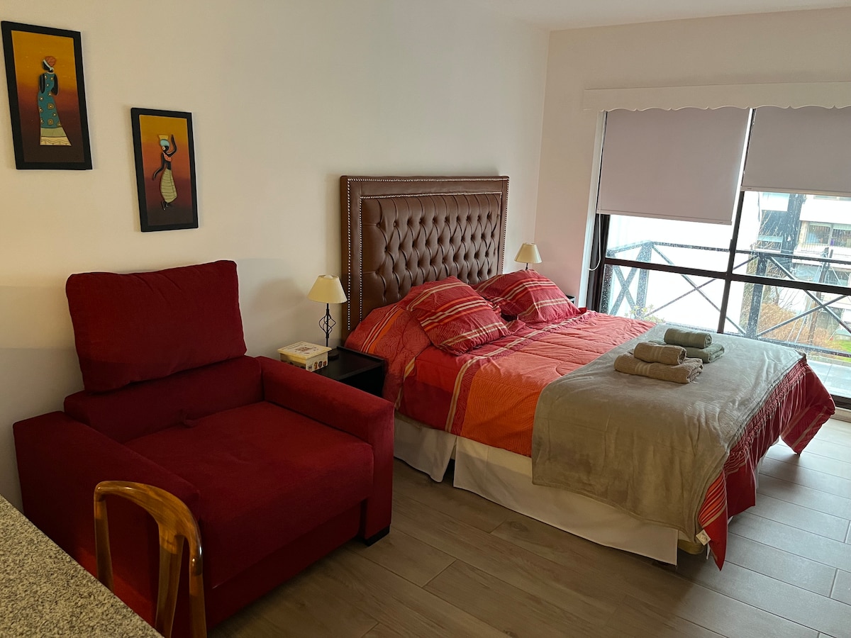 A cozy sleeping area features a spacious bed with a tufted headboard and soft bedding in warm tones. A red armchair is positioned nearby, complemented by bedside tables with lamps. Natural light filters through a large window, providing a bright and airy environment.