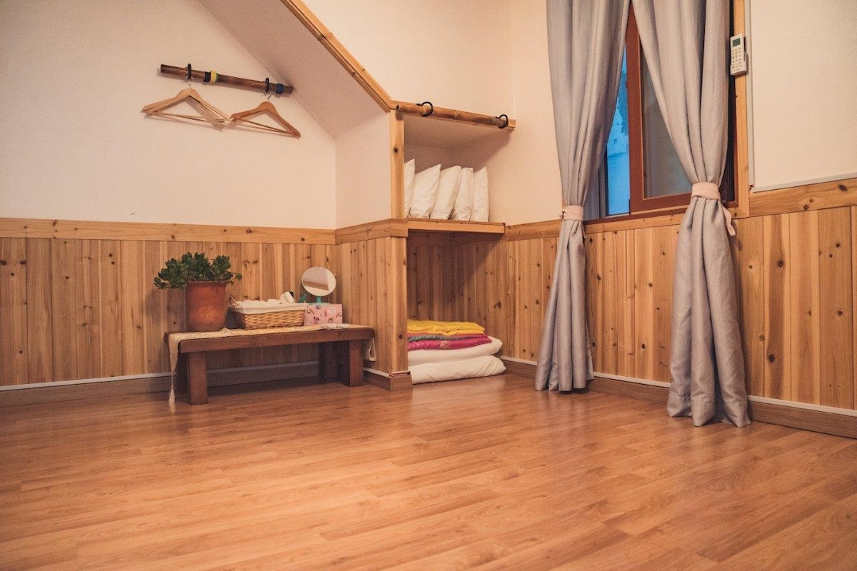 A cozy room is shown with wooden paneling along the lower walls. A bench offers neatly stacked linens, while a plant sits atop it. Light curtains frame a window, allowing glimpses of natural light to enhance the warmth of the wooden flooring.