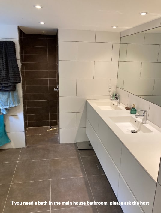The modern bathroom features a spacious dual vanity with a clean, white countertop. A large mirror reflects soft overhead lighting. The shower area, enclosed in glass, is visible to the left. Towel racks hold neat towels, contributing to the tidy appearance.