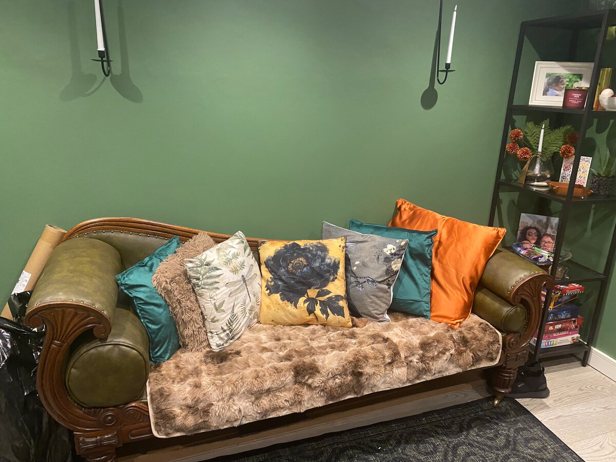 A stylish green wall serves as a backdrop for a vintage-style sofa adorned with an array of colorful pillows. A soft throw blankets one end, while a bookshelf nearby displays decorative items and small plants, adding character to the space.