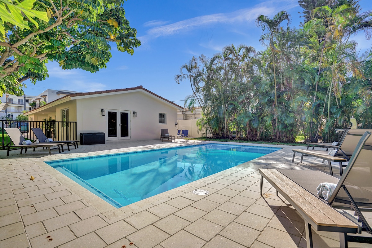 A resort-style pool is framed by tropical greenery, featuring a spacious deck with lounge chairs. The calm water reflects the blue sky, providing a serene setting for relaxation. A well-maintained patio creates an inviting atmosphere for outdoor enjoyment.