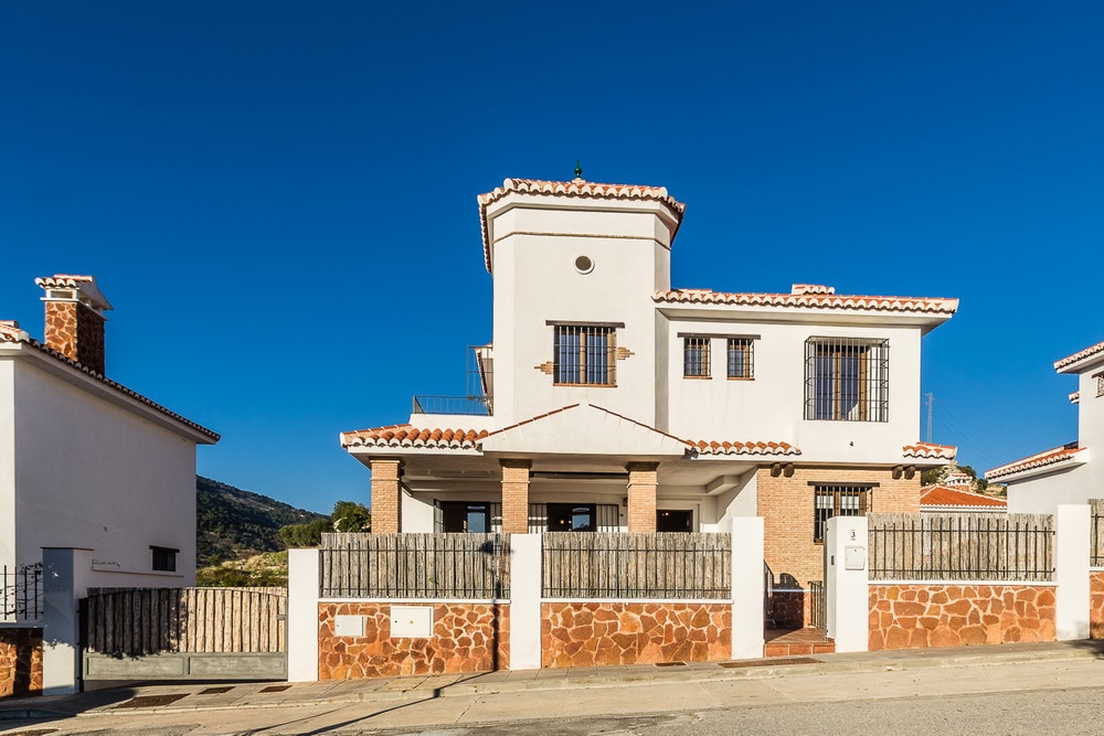 A villa with a modern architectural design is displayed against a clear blue sky. The two-story building features a combination of white stucco and brick elements. Large windows and a prominent turret add character, while a stone wall borders the property, ensuring privacy.