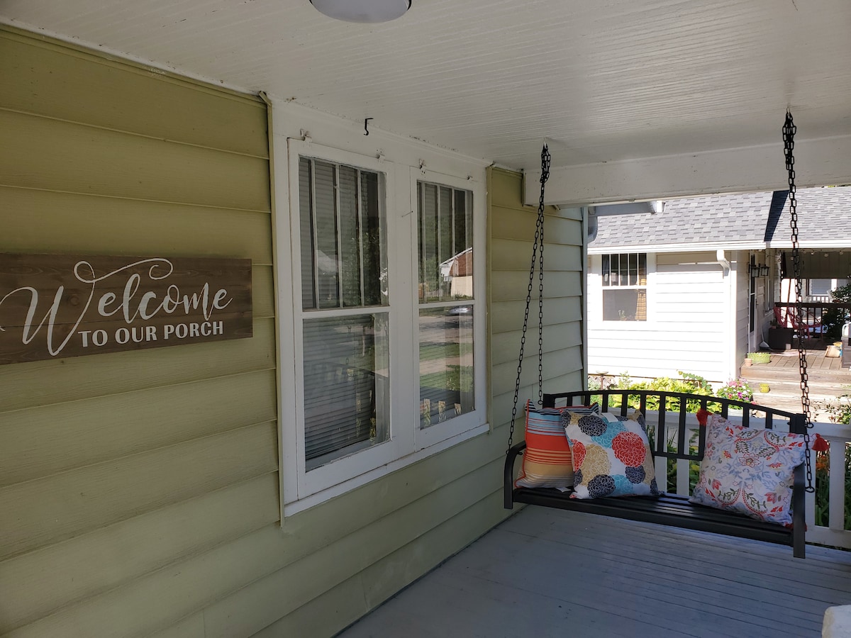 A front porch features a wooden swing adorned with bright, patterned cushions. Light-colored walls surround the porch, and a welcome sign is displayed nearby. Two windows allow natural light to enter, while views of the neighborhood can be seen in the background.