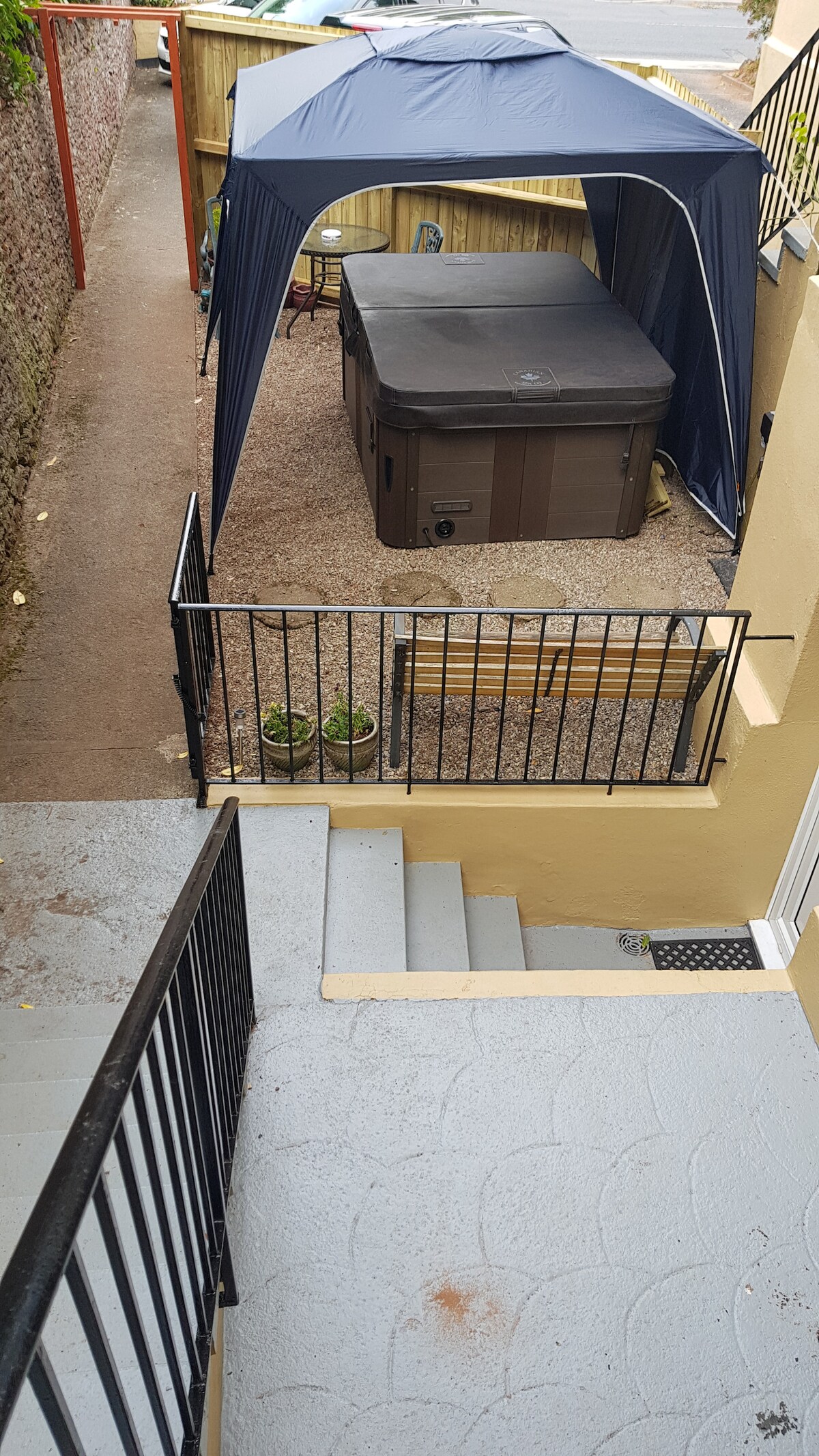 An outdoor area is featured, showing a private hot tub under a canopy. A bench is positioned nearby, with potted plants adding greenery. Stairs lead up to the entrance of the apartment, with a clean, textured surface visible in the foreground.