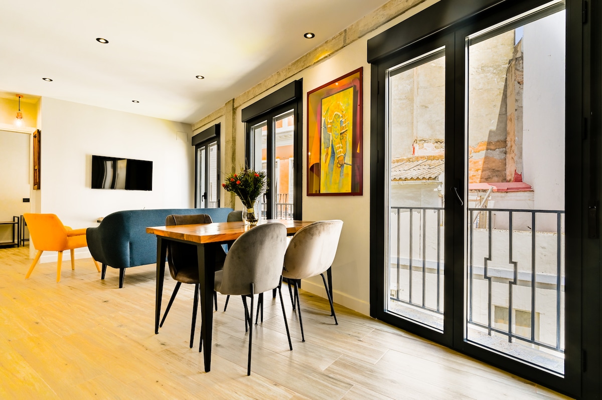 A dining area is presented with a wooden table surrounded by four comfortable chairs. A colorful artwork decorates the wall above the table. Large glass doors allow natural light to fill the space and provide views of the lively pedestrian street outside.