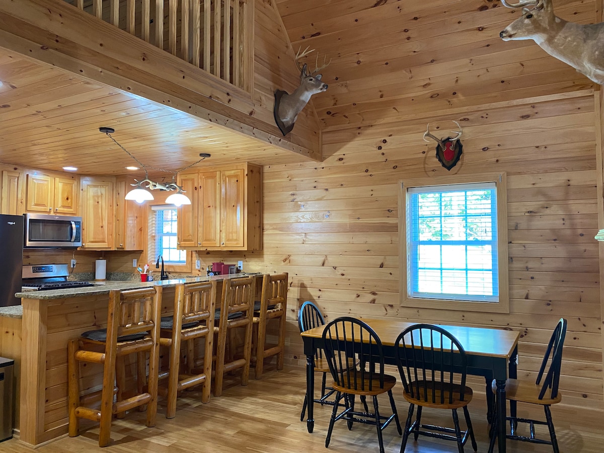 The kitchen and dining area features wooden walls and ceiling, complemented by natural light from a window. A spacious kitchen island with bar stools is visible, alongside a dining table surrounded by black chairs. Decor includes mounted deer heads, enhancing the rustic ambiance.