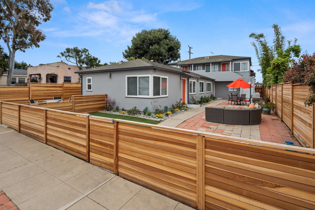 A charming exterior view showcases a remodeled condo surrounded by landscaped gardens and wooden fencing. A seating area with an orange umbrella is visible on the patio, and the pathway leads to additional outdoor spaces with vibrant greenery.