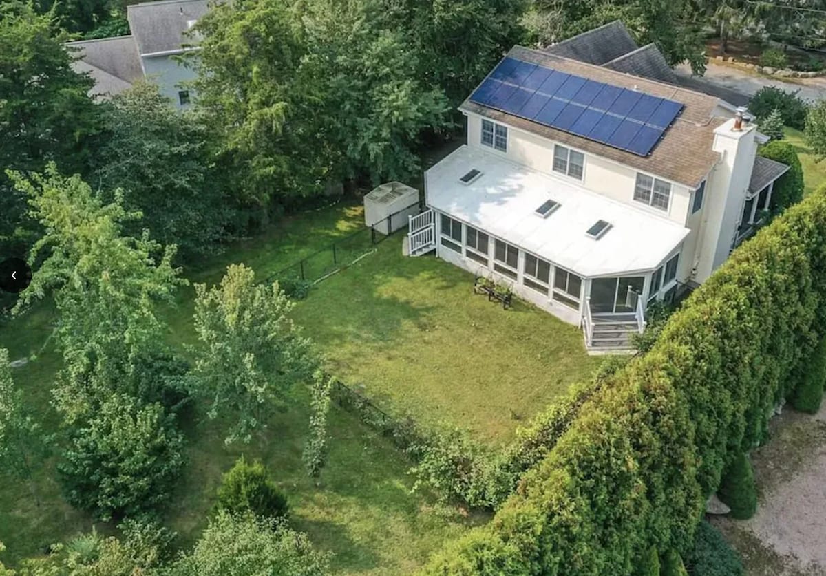 An aerial view showcases a spacious, well-maintained yard surrounding a two-story home. Solar panels are visible on the roof, while a large, climate-controlled sunroom extends from the back of the house. Lush greenery and tree line the property, enhancing the serene outdoor space.