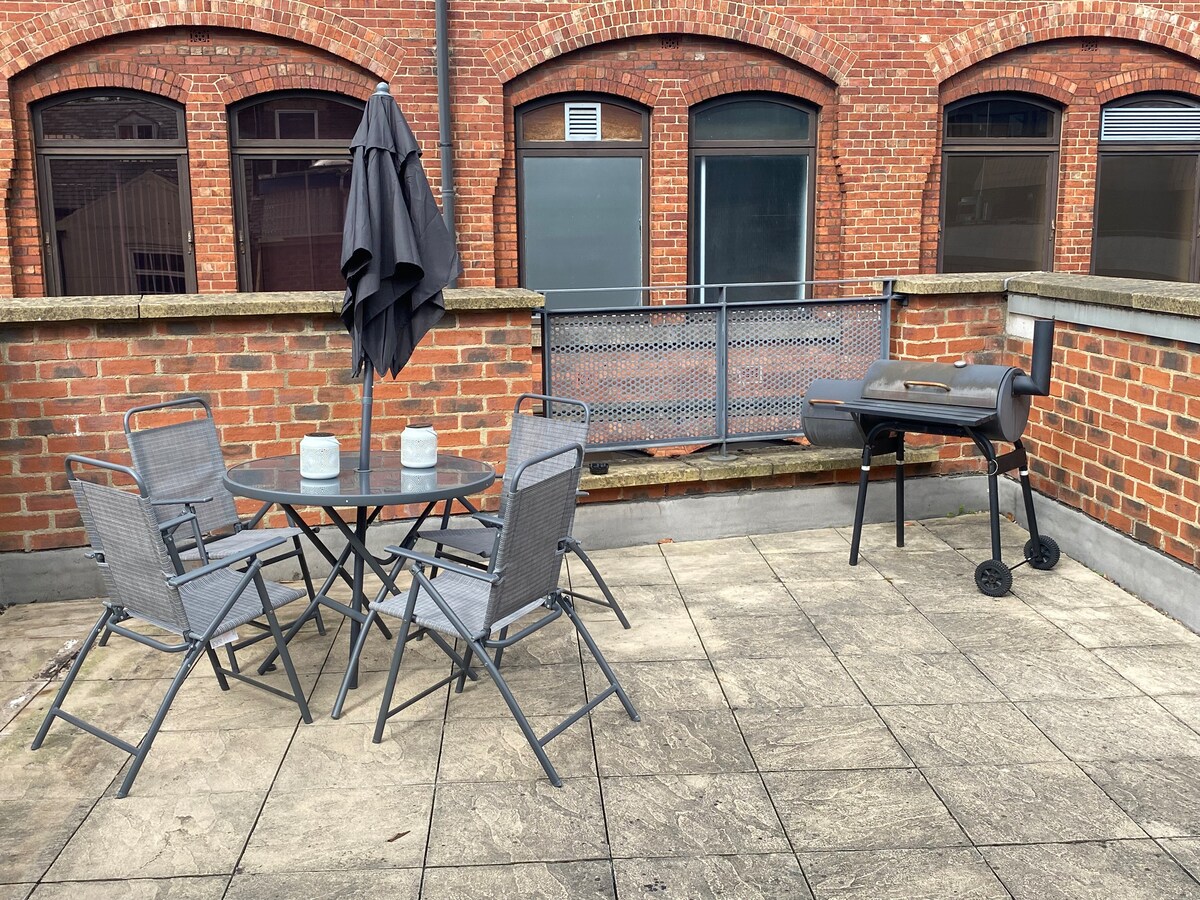 A private roof garden features a round table surrounded by four gray chairs, with two light-colored mugs placed on the table. A charcoal grill sits on a paved area, alongside a black umbrella mounted in a stand.