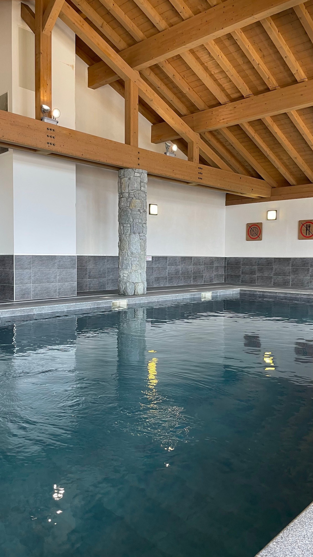 An indoor swimming pool features a tranquil blue surface reflecting light from above. The space is accented by wooden beams and a stone column, creating a relaxed atmosphere. Subtle artwork decorates the walls, enhancing the overall aesthetic of the area.