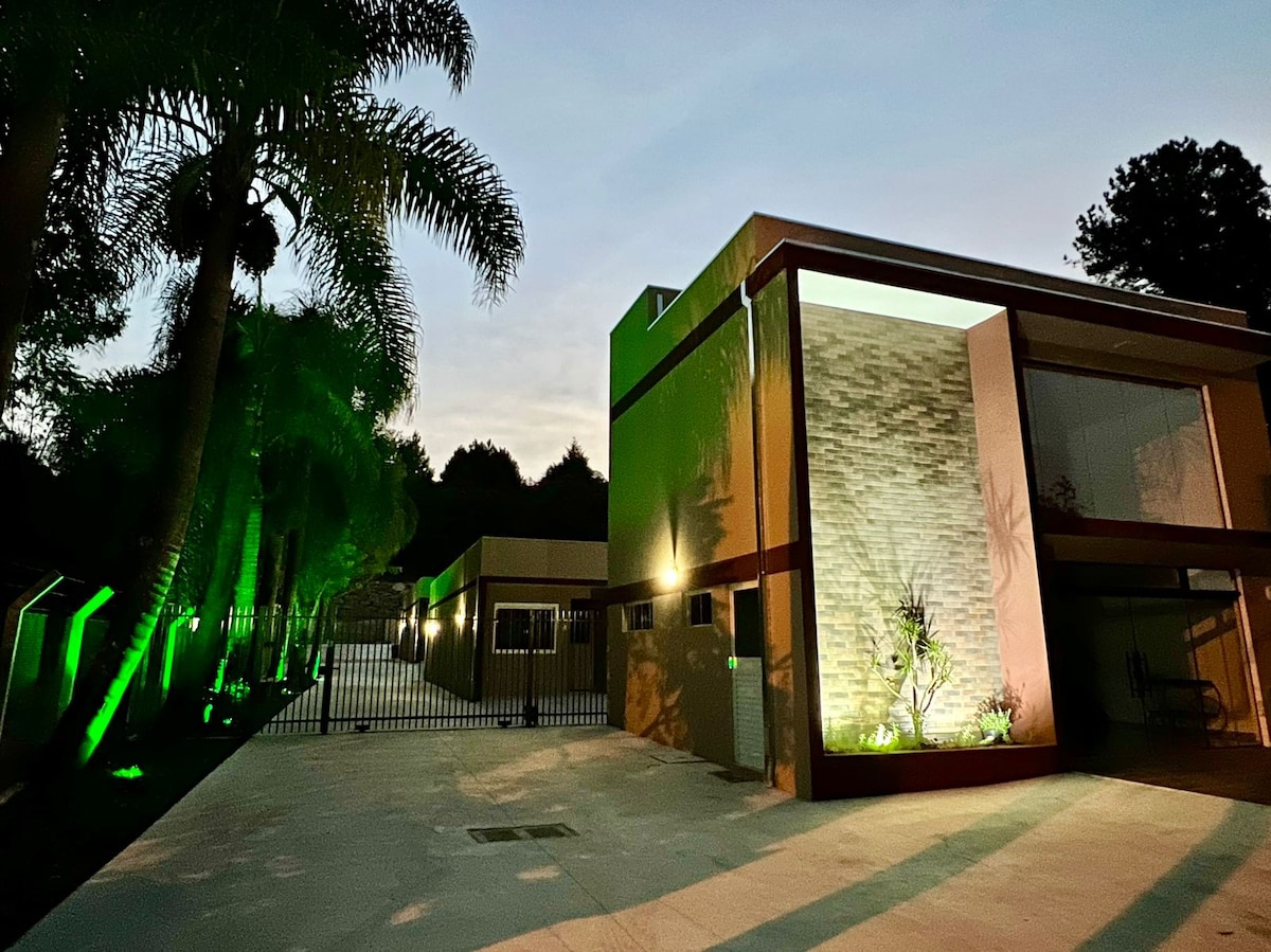 The exterior of the building features modern architecture with a combination of stone and green-painted walls. Soft lighting highlights the landscaping, including palm trees, while a spacious parking area is visible in the foreground. A secure fence surrounds the property, creating a sense of privacy.