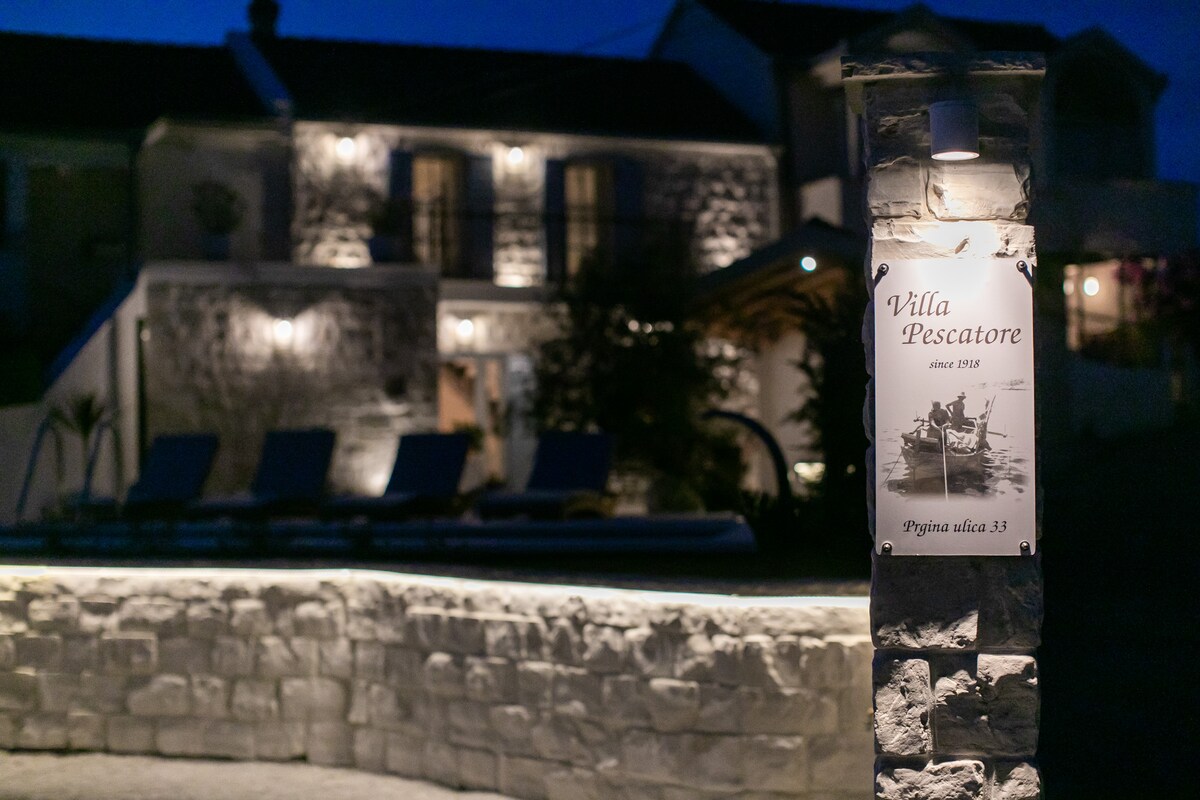The stone structure of Villa Pescatore is softly illuminated against the night sky, highlighting its rustic architecture. A welcoming sign displays the villa's name and history, while the surrounding area features cozy lighting, enhancing the ambience of the peaceful retreat.