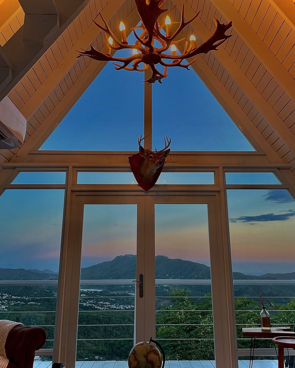 A striking triangular window offers a breathtaking view of the mountains and sky, framed by wooden walls. A rustic chandelier with antler accents hangs from the ceiling, and a decorative deer head adds a touch of alpine charm to the space.