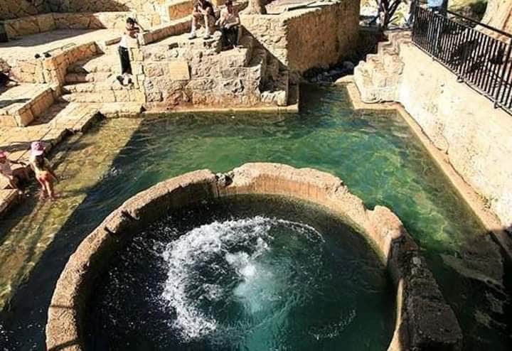 A natural spring is highlighted, with clear turquoise water flowing into a circular stone basin. Surrounding stone walls create tiered areas for seating, while visitors can be seen enjoying the tranquil environment. The setting showcases a blend of nature and architecture.