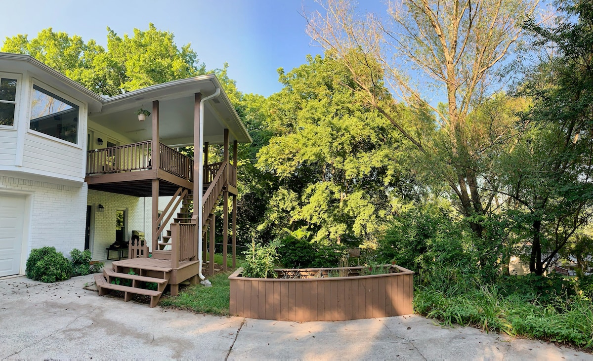 A private entrance leads up to the studio guest suite, bordered by lush greenery. A wooden deck includes seating, offering a serene space to enjoy the outdoors. A raised garden bed is visible, contributing to the natural setting of the property.