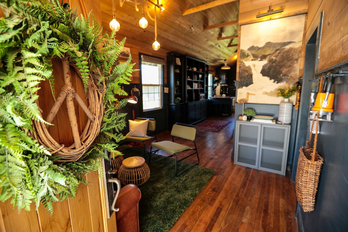 A welcoming entrance features a decorative wreath made of natural materials and lush greenery. The stylish hallway opens into a cozy living area, showcasing a mix of seating options, a wooden accent wall, and warm lighting fixtures, contributing to the home's inviting ambiance.