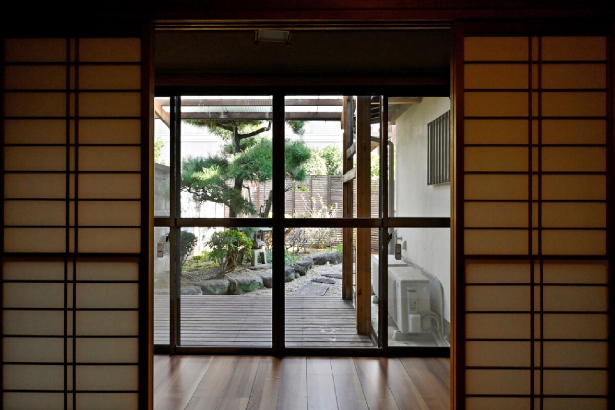 A spacious room is framed by sliding shoji doors, providing a view of a serene garden with a stone pathway and greenery. Natural light fills the space, highlighting the wooden flooring and creating a peaceful atmosphere.