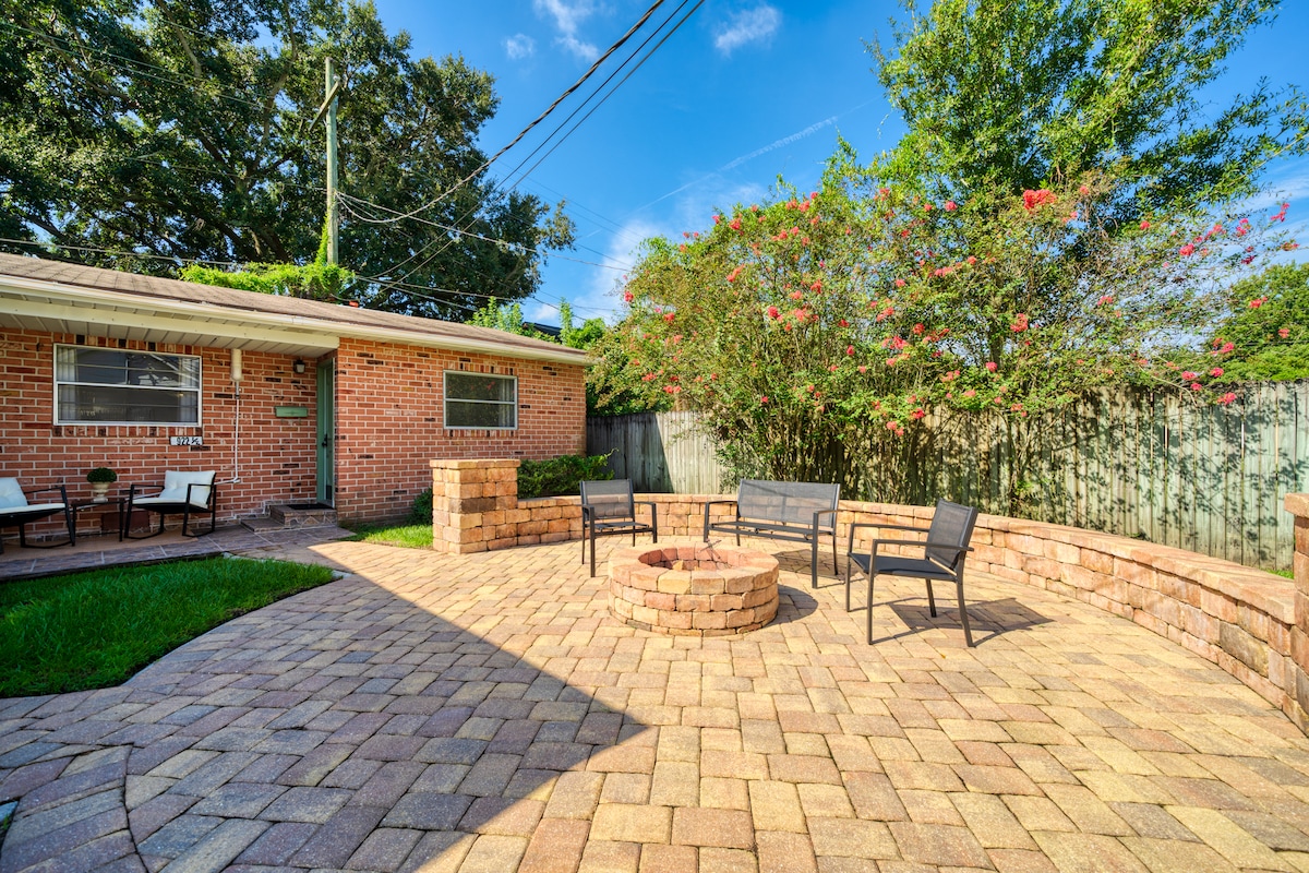 An outdoor patio is featured, with a circular paver fire pit surrounded by several black outdoor chairs. Lush greenery and blooming trees provide a natural backdrop, while a brick exterior of the guest suite and clear blue skies create a welcoming atmosphere.