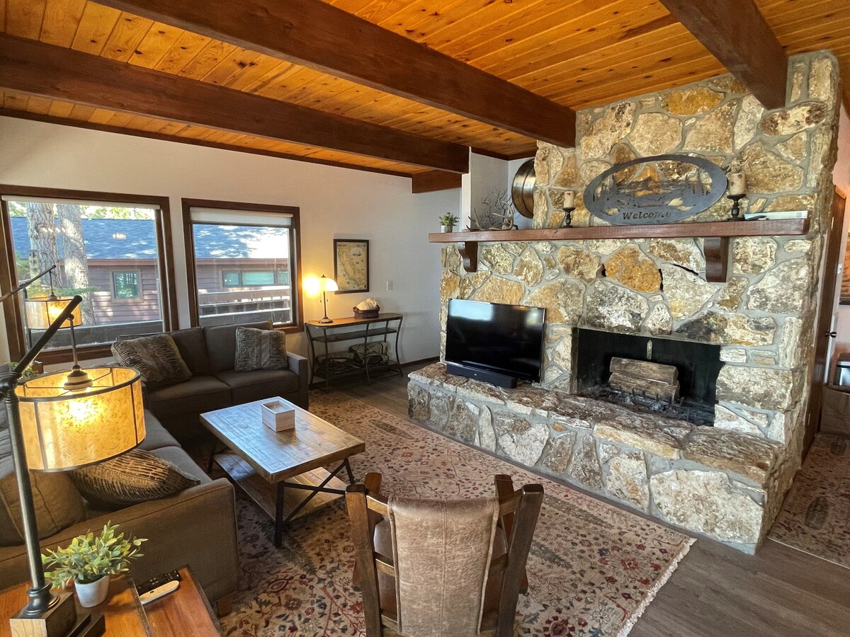 A cozy living area features a large stone fireplace and wooden beams above. Comfortable seating includes a plush sofa and a rustic dining table. Natural light streams through the windows, enhancing the inviting atmosphere of the space.
