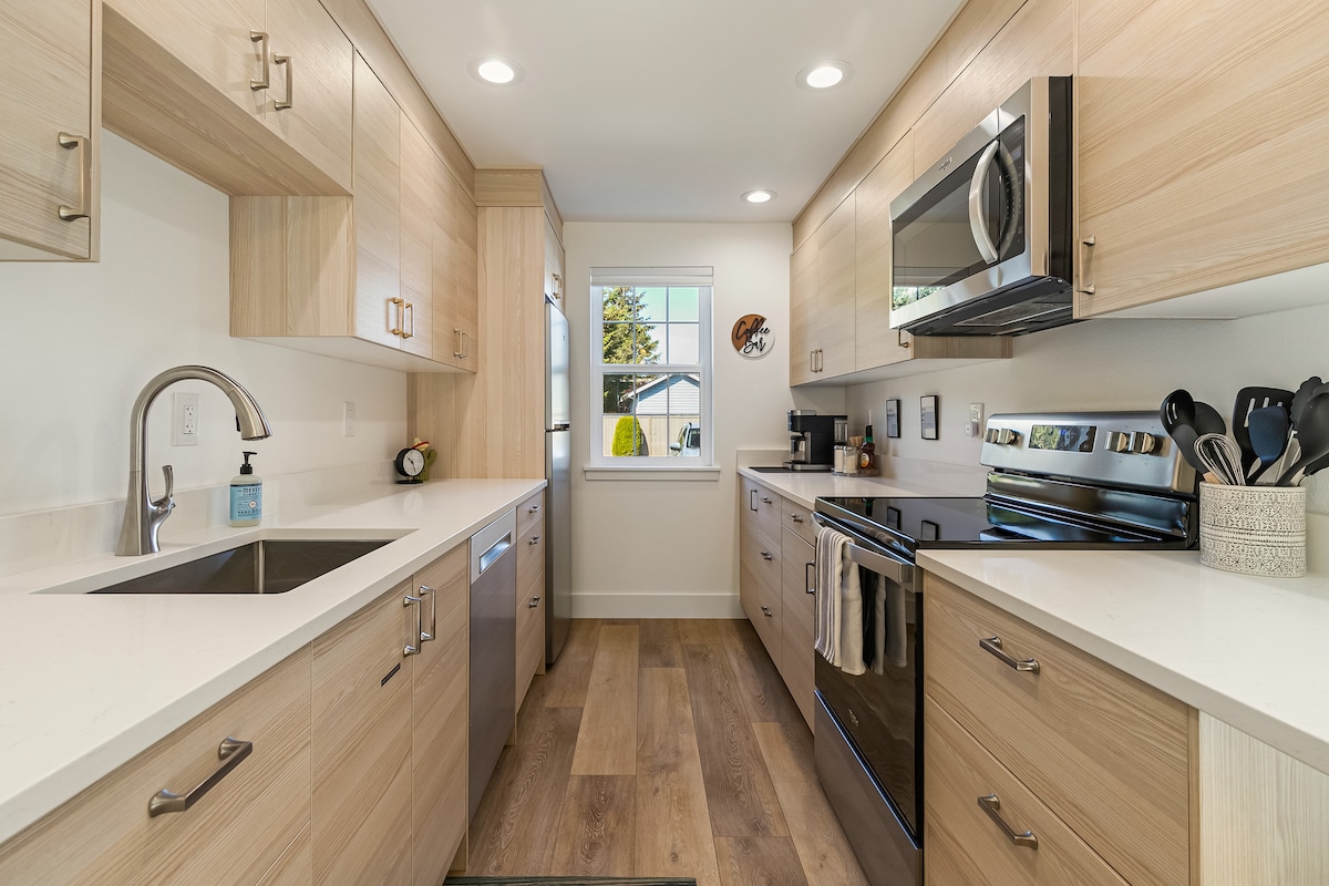 A modern kitchen is presented with light wood cabinetry and stainless steel appliances. The space features a double sink, an electric oven, and a microwave. A window allows natural light to illuminate the countertop area, which is equipped with cooking utensils and a small decorative item.
