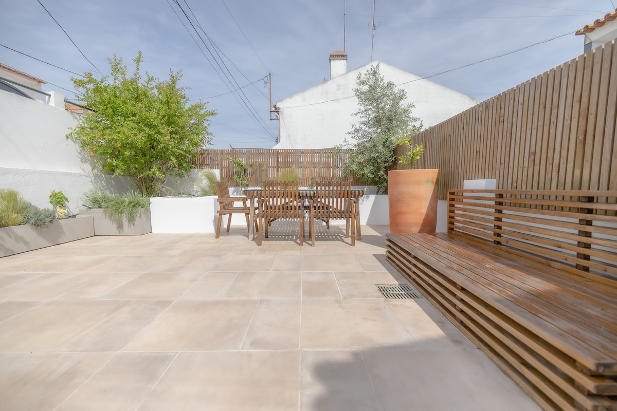 The outdoor terrace features a spacious area with a wooden dining table and matching benches, surrounded by greenery. Minimalistic landscaping includes planters with various plants, while a wooden fence provides privacy, enhancing the peaceful atmosphere of the space.
