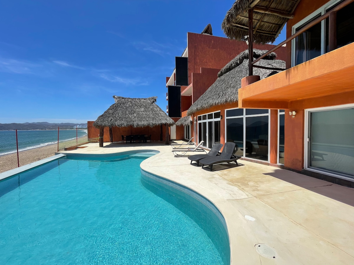 The outdoor area features a curvy pool surrounded by a sun deck with lounge chairs. Thatched-roof palapas provide shaded seating. The backdrop shows a vibrant orange and red villa with extensive glass windows, offering views of the nearby beach and ocean.