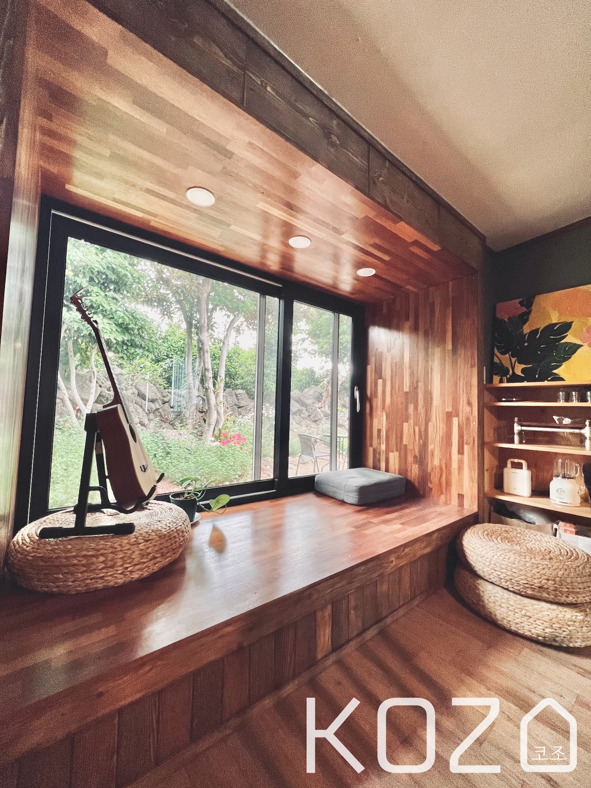 A cozy nook is featured with a large window offering views of the garden. Natural wood paneling adds warmth. The space includes cushions on a wooden ledge and braided baskets on the floor, enhancing a sense of relaxation and comfort.