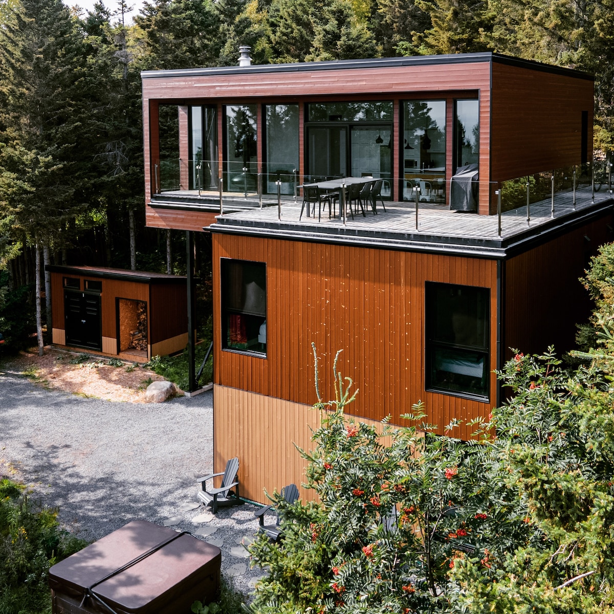 A modern chalet is nestled among trees, featuring a two-story design with large glass windows. A spacious balcony is visible, furnished with a dining table and chairs. The exterior is clad in wood, complementing the natural surroundings.