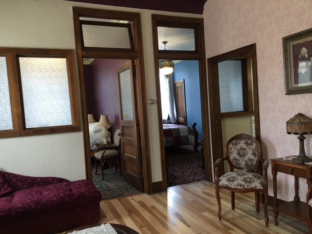 The suite entrance is framed by elegant wooden doors, leading into a spacious living area with rich hardwood flooring. A plush, deep-colored seating area is visible, alongside a decorative side table and a vintage lamp. The adjoining bedroom features a colorful wall and is softly illuminated by natural light.