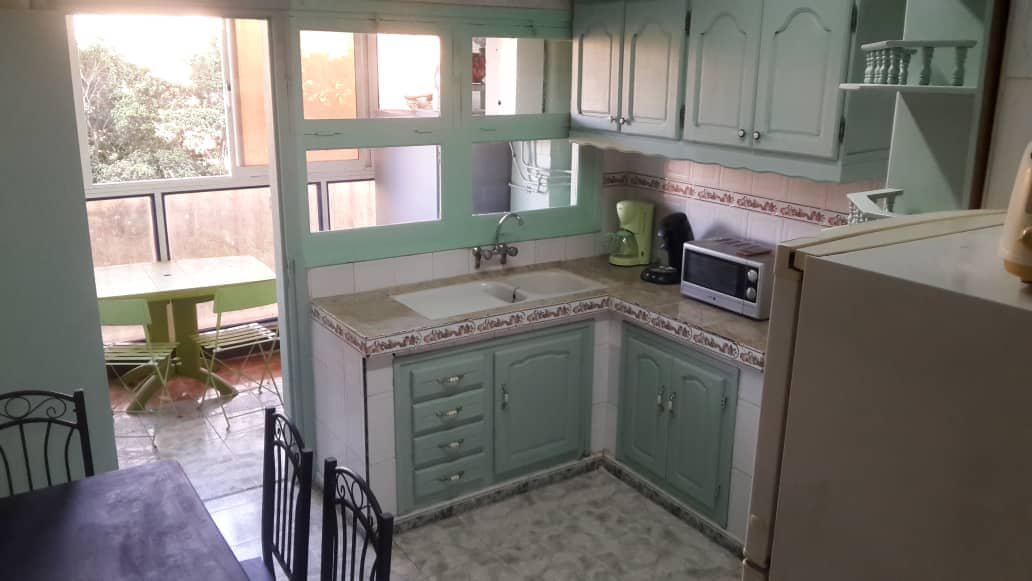 A functional kitchen features light green cabinetry with multi-panel windows allowing natural light. Essential appliances, including a refrigerator, microwave, and coffee maker, are present. A small dining table is situated next to the window, complementing the room's layout. The area is organized and inviting.