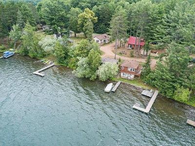 An aerial view captures the peaceful waterfront cabin situated among lush trees along Big Arbor Vitae Lake. Multiple docks extend into the water, providing access for boating. Surrounding greenery and a gravel path lead to nearby buildings, harmonizing with the natural landscape.